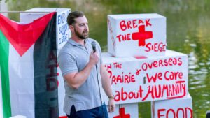 A white man stands with a microphone in front of boxes that says "Break the Siege" and "In the prairie and overseas, we care about humanity." There is also a Palestinian flag and a lake behind the boxes.