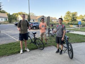 3 young men stand with each other in front of a parking lot on grass.