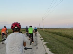 Several cyclists head down a road with corn fields visible and the moon