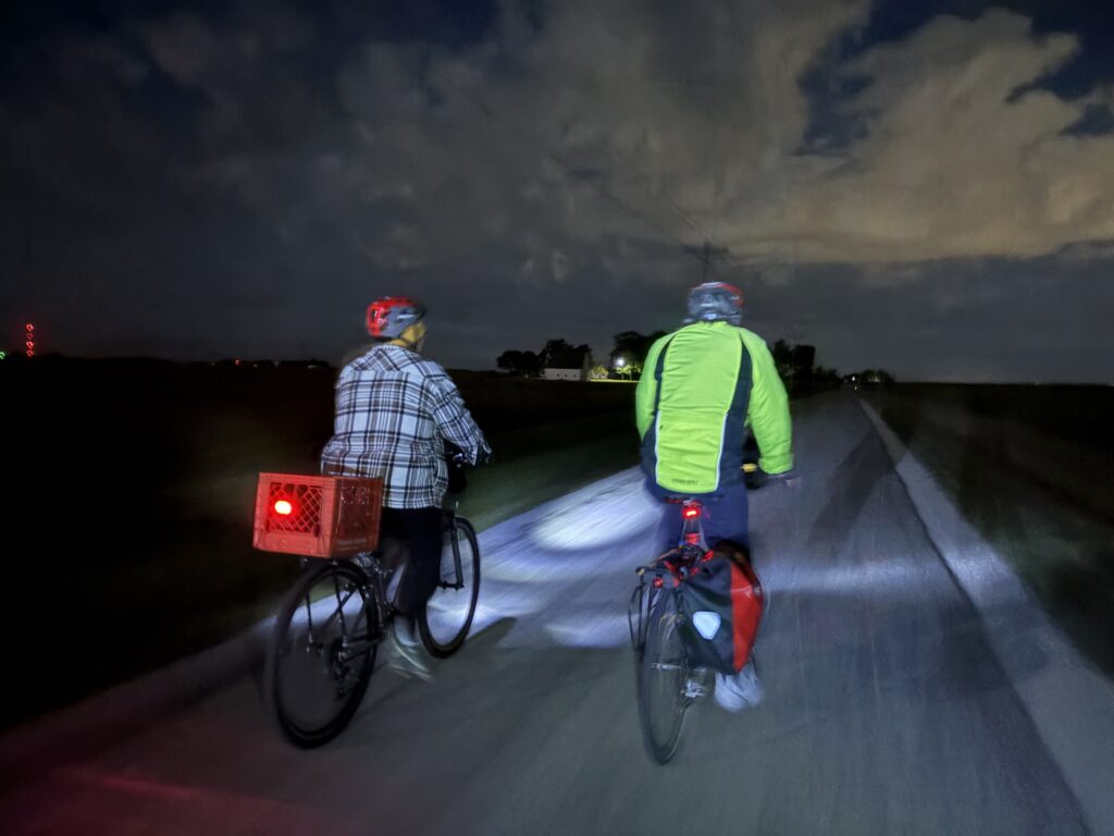 Two cyclists ride in the dark, lights on in front of them.
