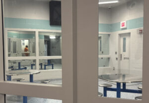 A white room with overhead lights and a few metal tables seen through a glass door. A person in orange clothes is visibile through the room's windows across the hallway.