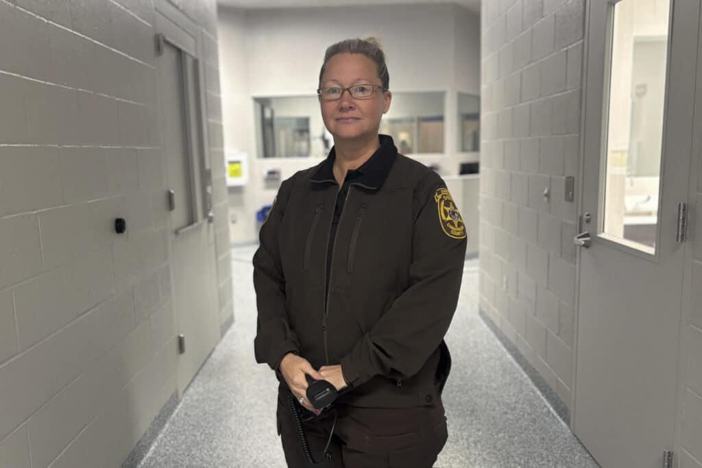 A woman with glasses in a brown police uniform stands in a white hallway.