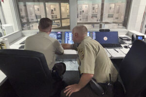 Two men in police uniforms sit in chairs in front of multiple monitors. One man leans over to the other and gestures to something on a monitor.