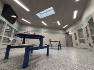 A low angle shows two metal tables in a white room with windows to hallways. A small skylight is in the middle of the ceiling surrounded by several overhead lights.