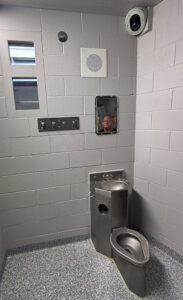 A room with white brick walls and a small window high up on the wall. A security camera sits in the upper right corner of the room, above a metal toilet and a mirror hanging on the wall, in which a woman's reflection is visible.