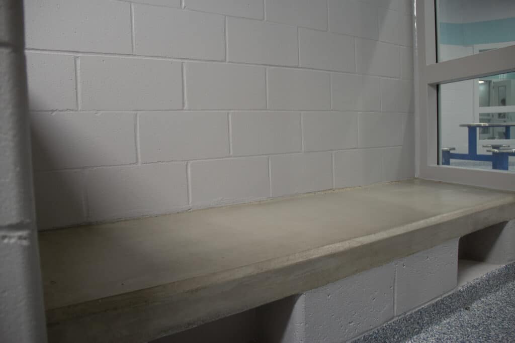 A bare concrete slab is on one side of a white brick room. Metal tables can be seen through a window next to the slab.