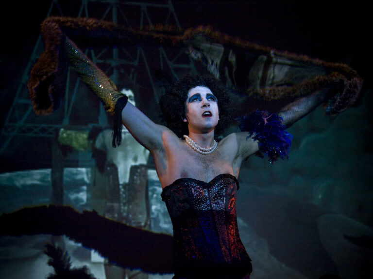 A person in white face paint and a dark corset stands in front of a screen projecting a scene from "The Rocky Horror Picture Show"