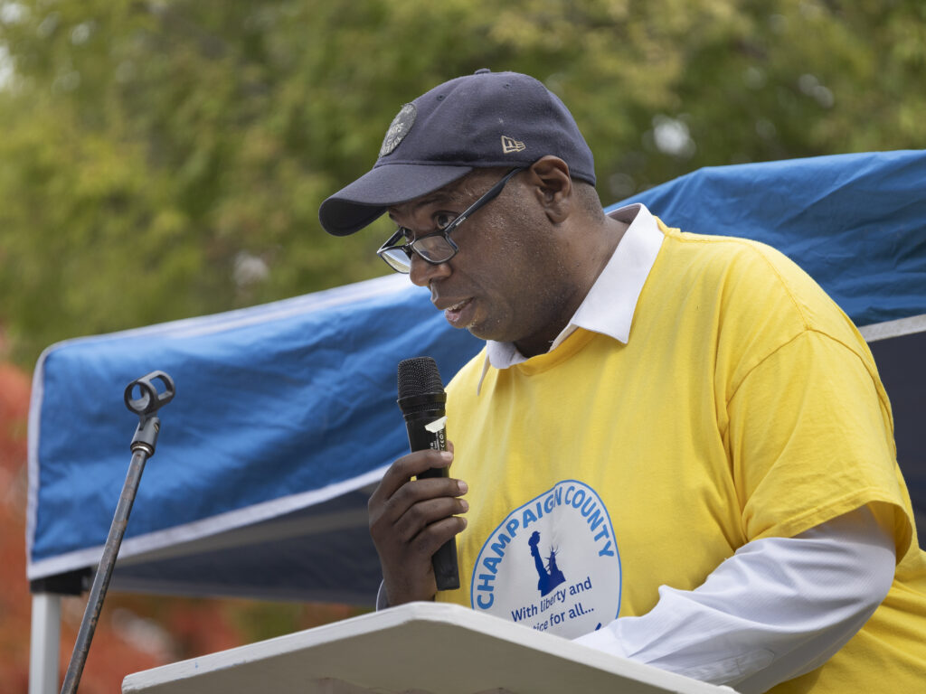 A black man in a baseball cap and a yellow shirt with an organization logo on it speaks into a microphone at a podium outside.