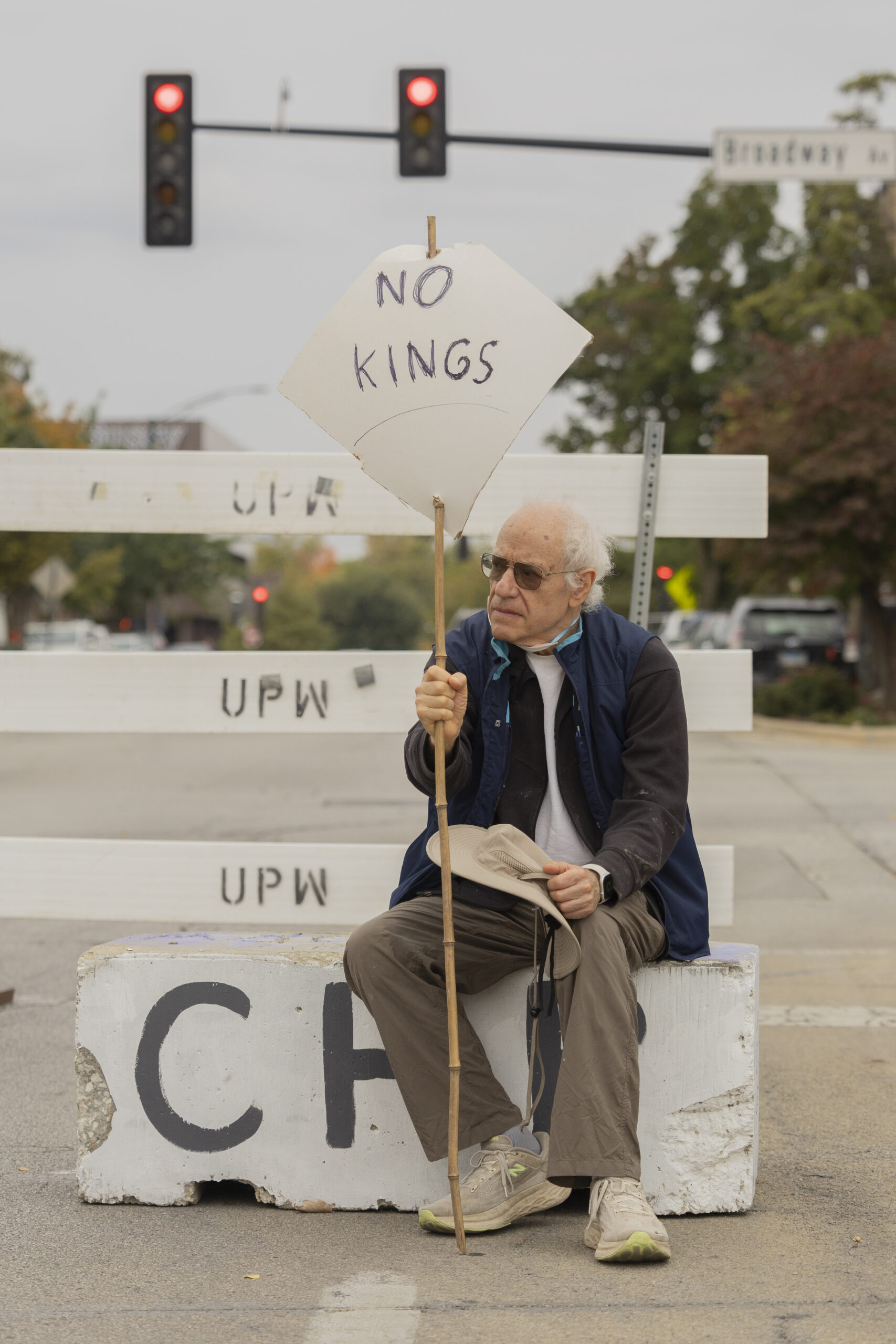 A man sits on a concrete baricade holding a sign that reads "No Kings" on one side.