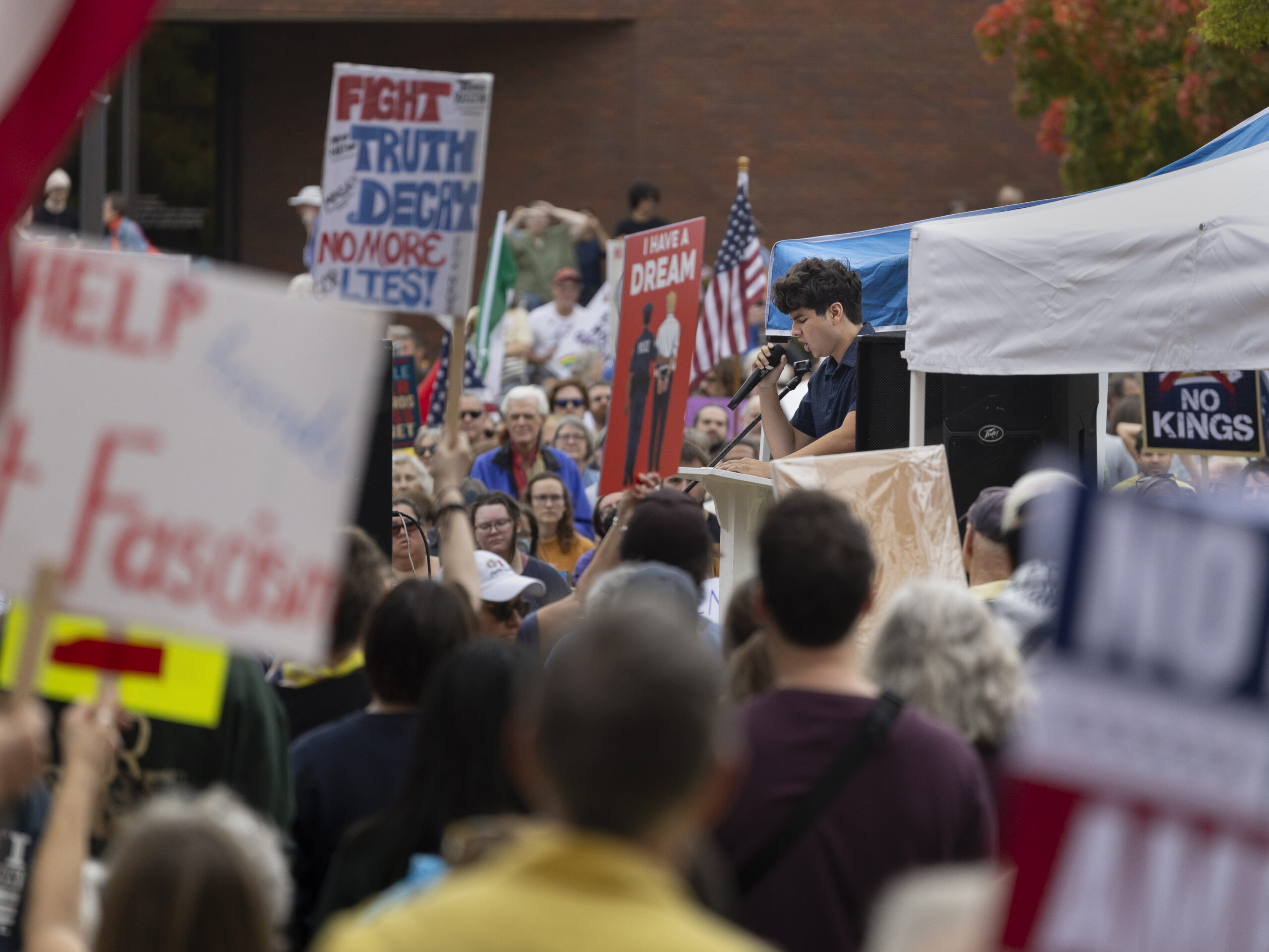A young man holds a microphone and speaks to a dense crowd, some of whom hold signs or flags.