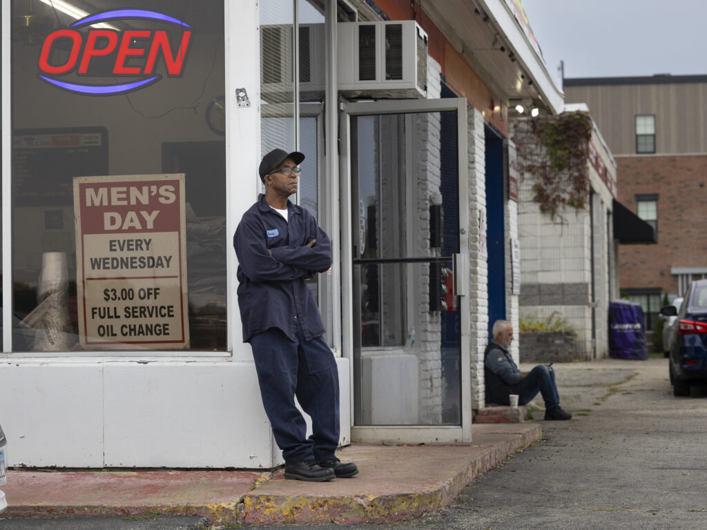 A man in a mechanics uniform leans against the side of his shop. An open sign glows in the window and another man sits on the sidewalk along the front of the building.