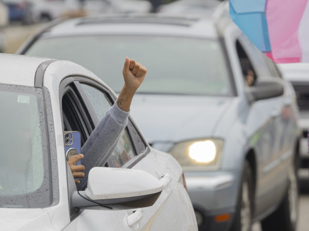 A person sitting in the driver's seat of a car raises their fist out of the window and films with their phone.