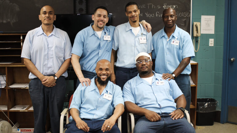 Six men in blue polos lean on one another and smile in a classroom.
