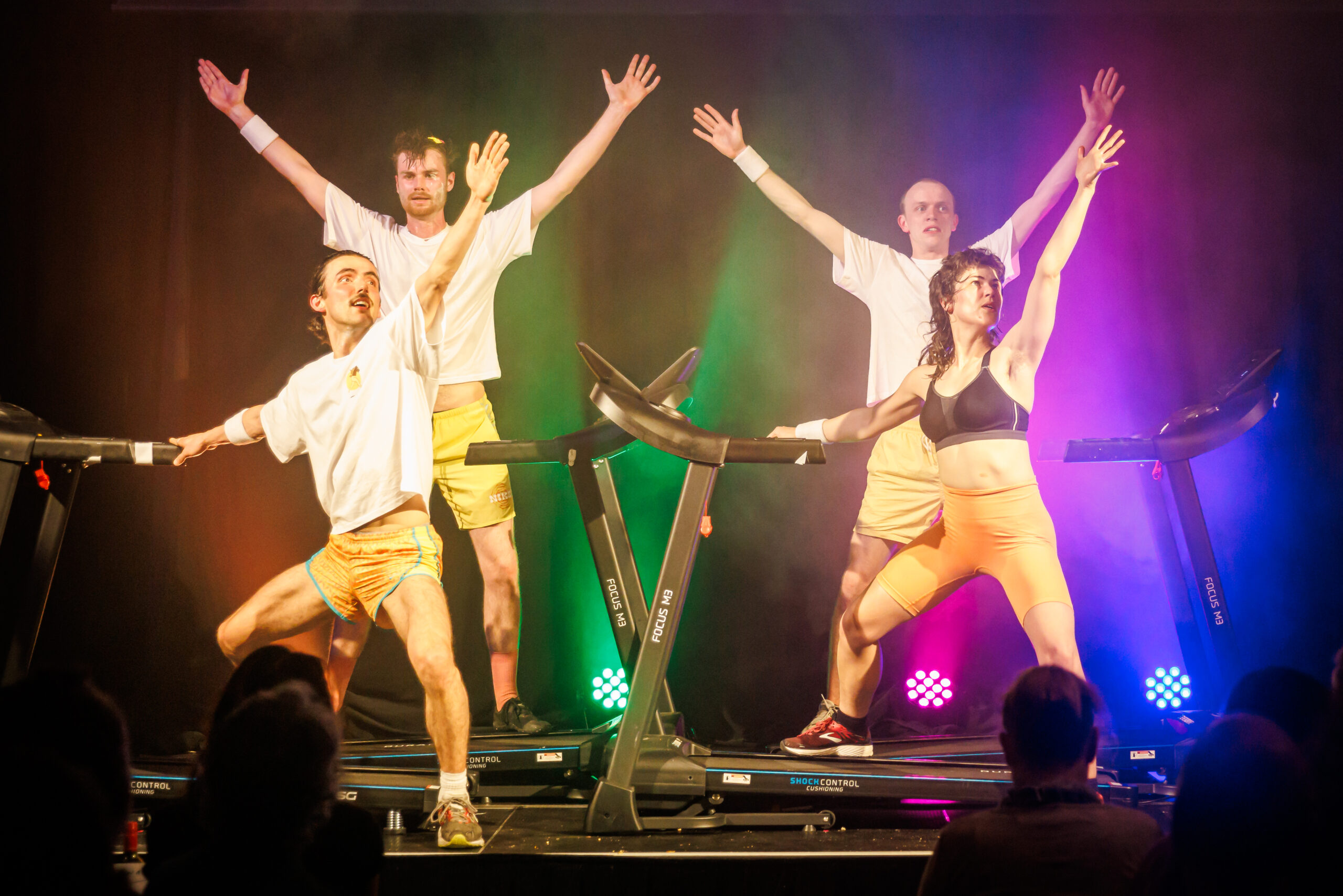 Four performers stand and pose on treadmills on a stage.