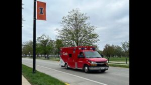 A red Carle Arrow Ambulance drives down a road with a University of Illinois banner hanging from a nearby lightpole.