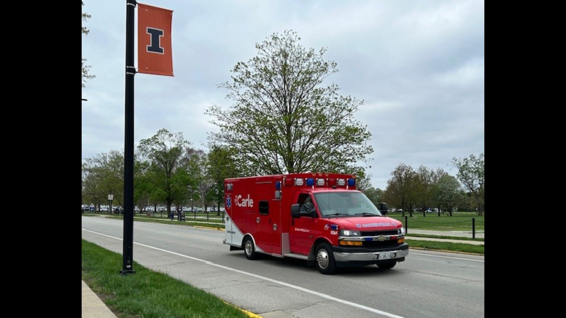 A red Carle Arrow Ambulance drives down a road with a University of Illinois banner hanging from a nearby lightpole.