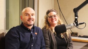 A white man and a white woman smile in a radio studio. There's a microphone in the foreground.