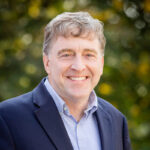 A white man in a suit smiles for a portrait outside.
