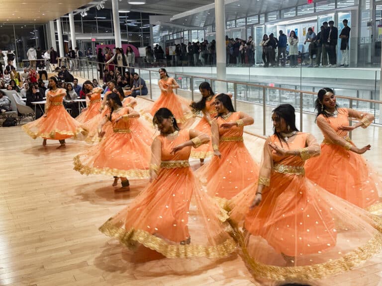 Several women in peach-colored dresses with gold trim dance together in a large open room with wood floors.