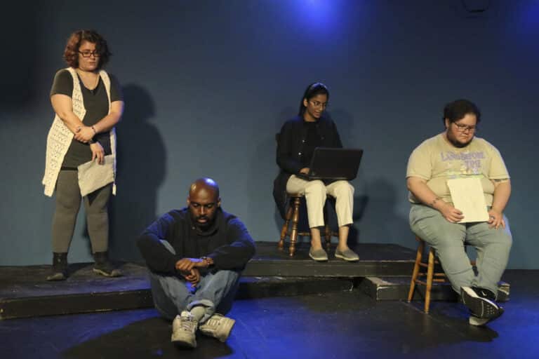 Four people in neutral clothing stand or sit on a small black stage lit with dim blue lights.