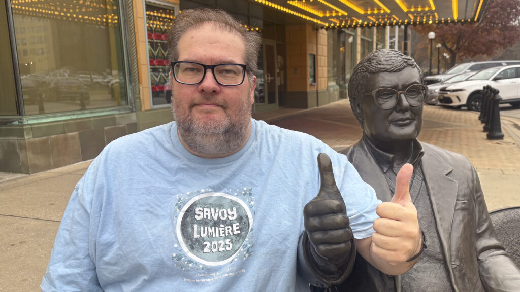 A man in a blue t-shirt reading "Savoy Lumiere 2025" sits next to a statue of film critic Roger Ebert on a bench.