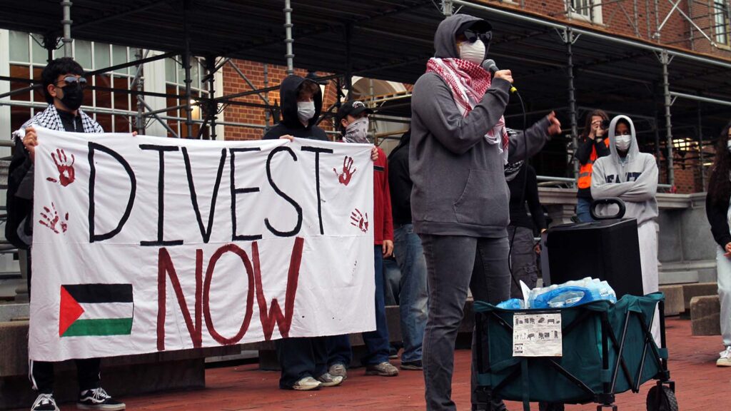 A person in a mask speaks into a microphone in front of a "Divest Now" banner.