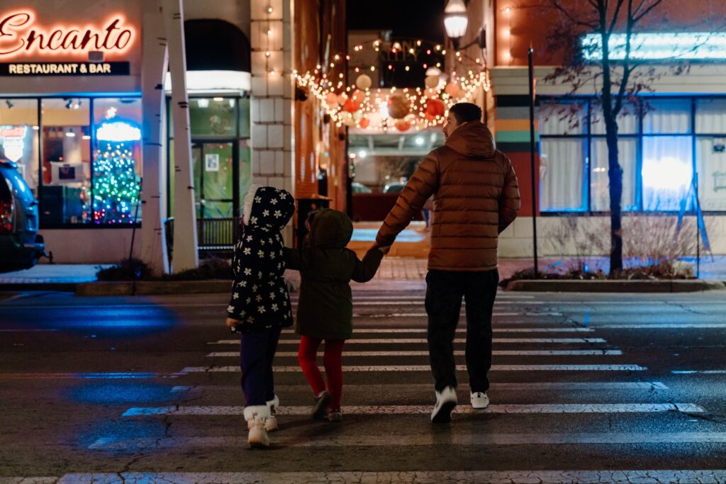 An adult and two children cross a road toward a lighted alleyway.