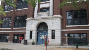A brick school building with an arched doorway and white columns is in the background. In the foreground is a "Keep Out" sign and rope cordoning off the entrance. This is Decatur's 100-year-old Dennis Lab School in 2023 after it was deemed unsafe for students.