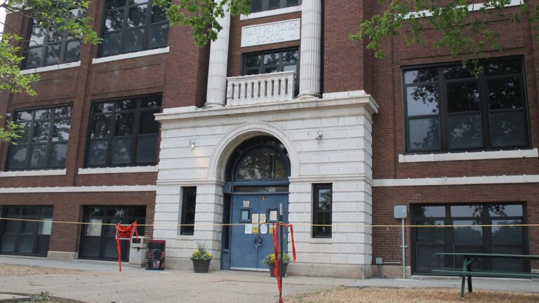 A brick school building with an arched doorway and white columns is in the background. In the foreground is a "Keep Out" sign and rope cordoning off the entrance. This is Decatur's 100-year-old Dennis Lab School in 2023 after it was deemed unsafe for students.