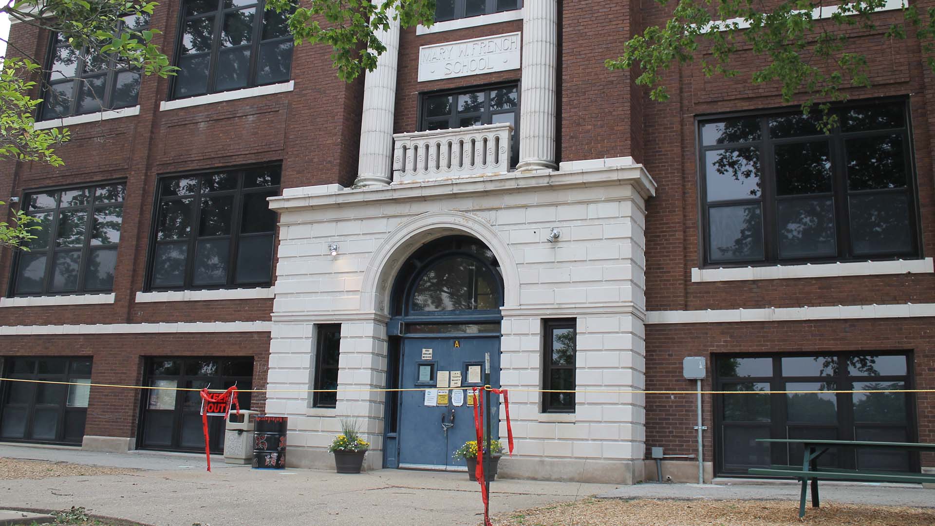 A brick school building with an arched doorway and white columns is in the background. In the foreground is a "Keep Out" sign and rope cordoning off the entrance. This is Decatur's 100-year-old Dennis Lab School in 2023 after it was deemed unsafe for students.