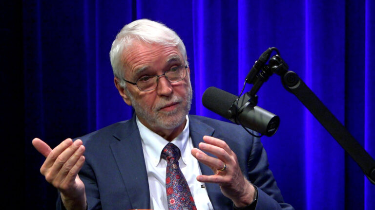 A white-haired man in a suit gestures with his hands, in front of a blue curtain. He is speaking into a studio microphone.