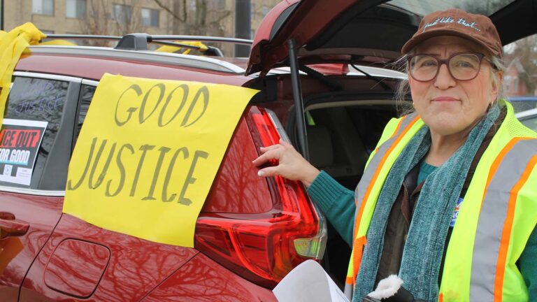 A white woman in a yellow vest stands by a car with a yellow sign that says "Good Justice."