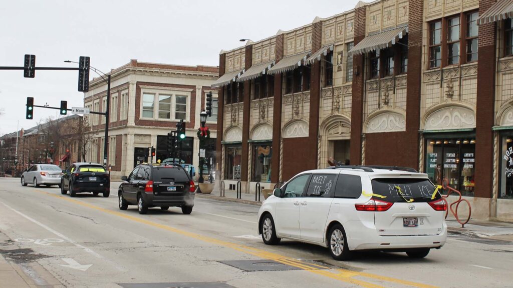 Four cars wait in a turn lane in downtown Urbana. The rear car has "Melt ICE" written on the windows and yellow streamers.