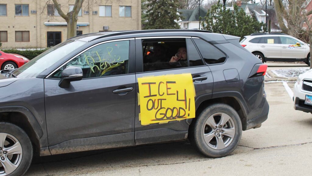A passenger rolls down the car window to blow a noisemaker. A yellow sign on the car reads "ICE OUT 4 GOOD!" and writing on another window reads "RIP Renee."