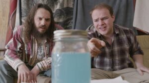 Two men sitting on a couch start at a jar full of light blue liquid.