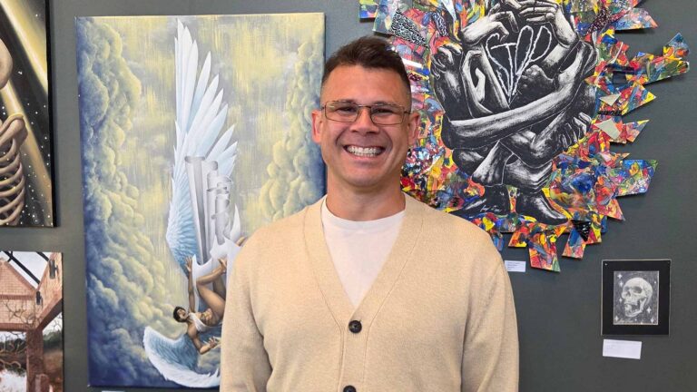 A white man with glasses, a beige sweater and a smile stands in front of an art exhibit.