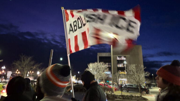 A group of people in winter clothes walk outside at night. One person waves a flag reading "Abolish ICE" above their head.