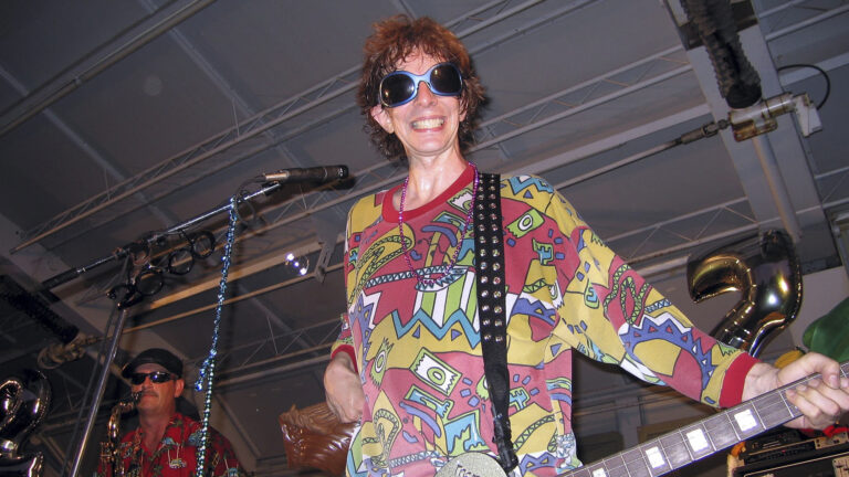 A man in a brightly colored smiles while holding a guitar and standing in front of a microphone.