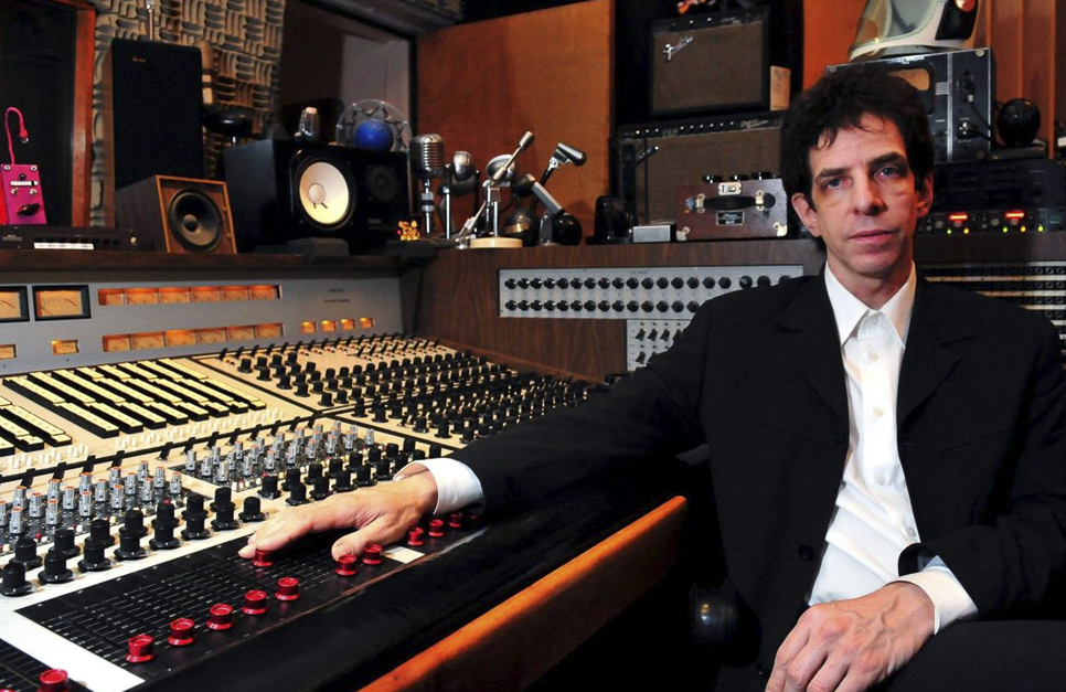 A man in a white collared shirt and black blazer sits next to a large sound board in a recording studio.