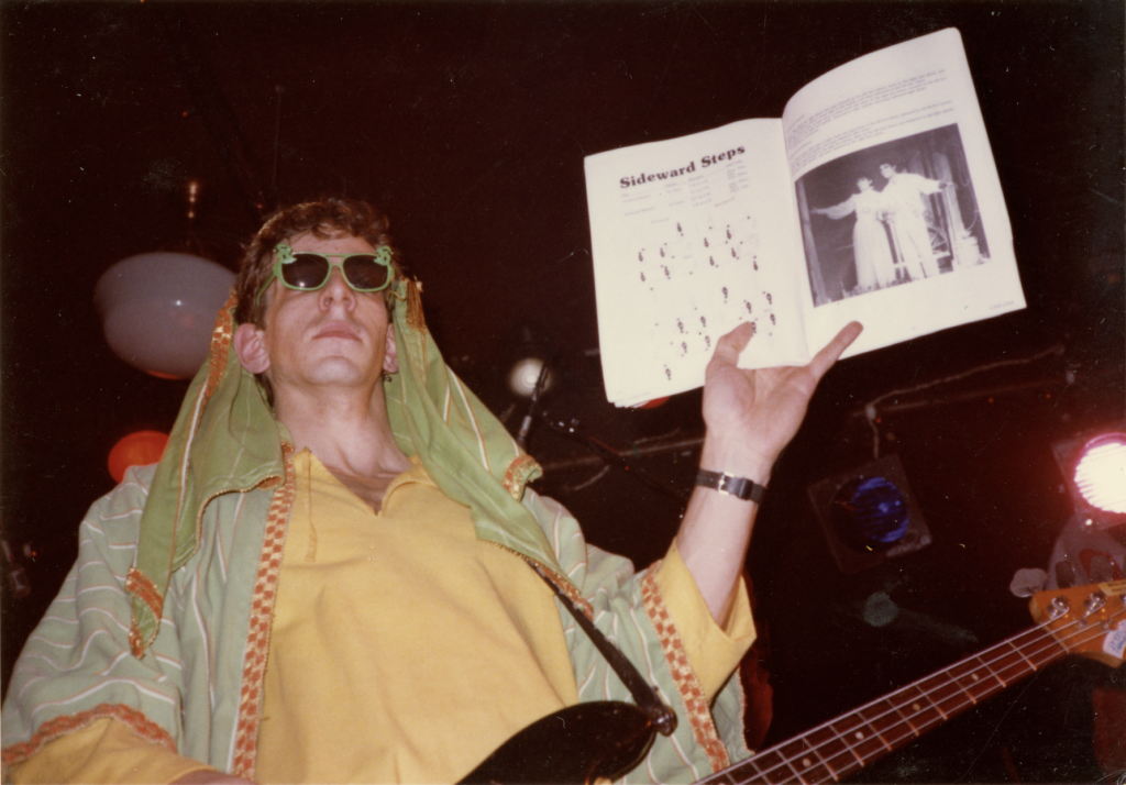A man in bright clothing plays the bass guitar with one hand while holding a book in the other.
