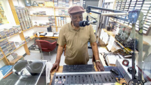 An older Black man wearing a cap stands in front of a sound board and speaks into a microphone in a small studio.