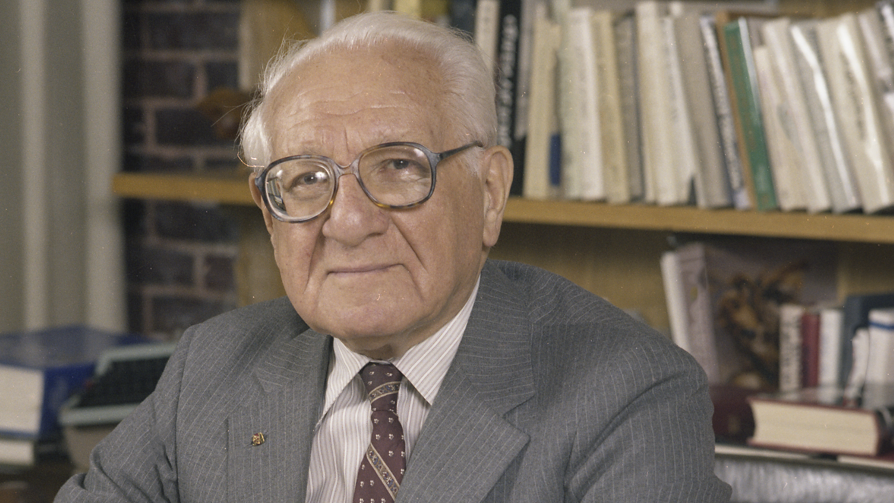 An older man wearing glasses and a grey suit softly smiles at the camera. Behind him are shelves full of books.