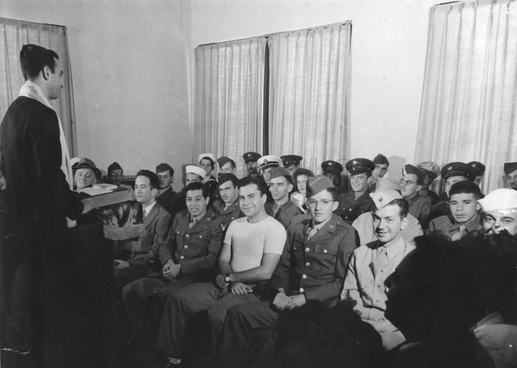 A black and white photograph of soldiers and sailors sitting in a meeting room while a man speaks in front of them.