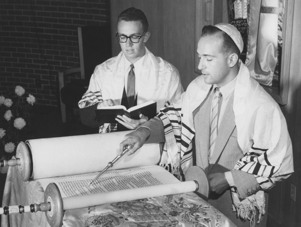 A black and white photo of two men using a yad to read the Torah.