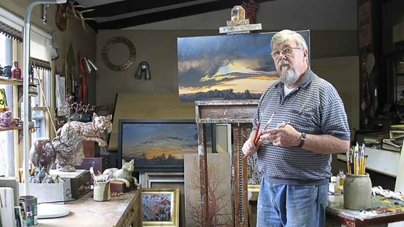 A man stands in front of an easel with a painting on it in a room full of similar paintings.