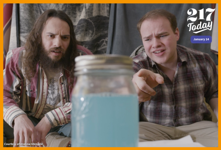 Two men sitting on a couch look at a jar filled with blue liquid.