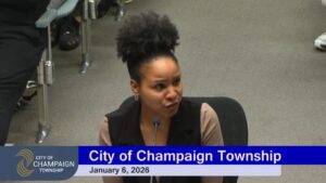 A screenshot showing a seated woman speaking into a microphone as a digital banner below her reads "City of Champaign Township January 6, 2026"