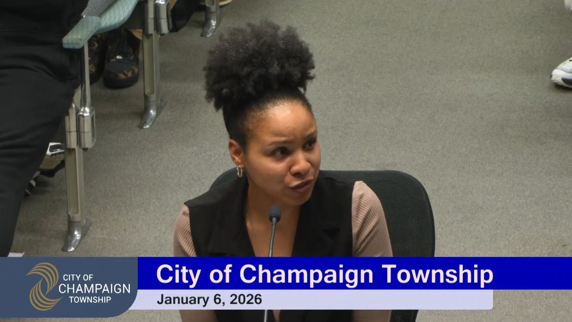 A screenshot showing a seated woman speaking into a microphone as a digital banner below her reads "City of Champaign Township January 6, 2026"