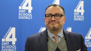 A man with cglasses, a suit and a colorful tie smiles at someone off camera.