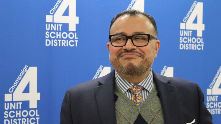 A man with cglasses, a suit and a colorful tie smiles at someone off camera.